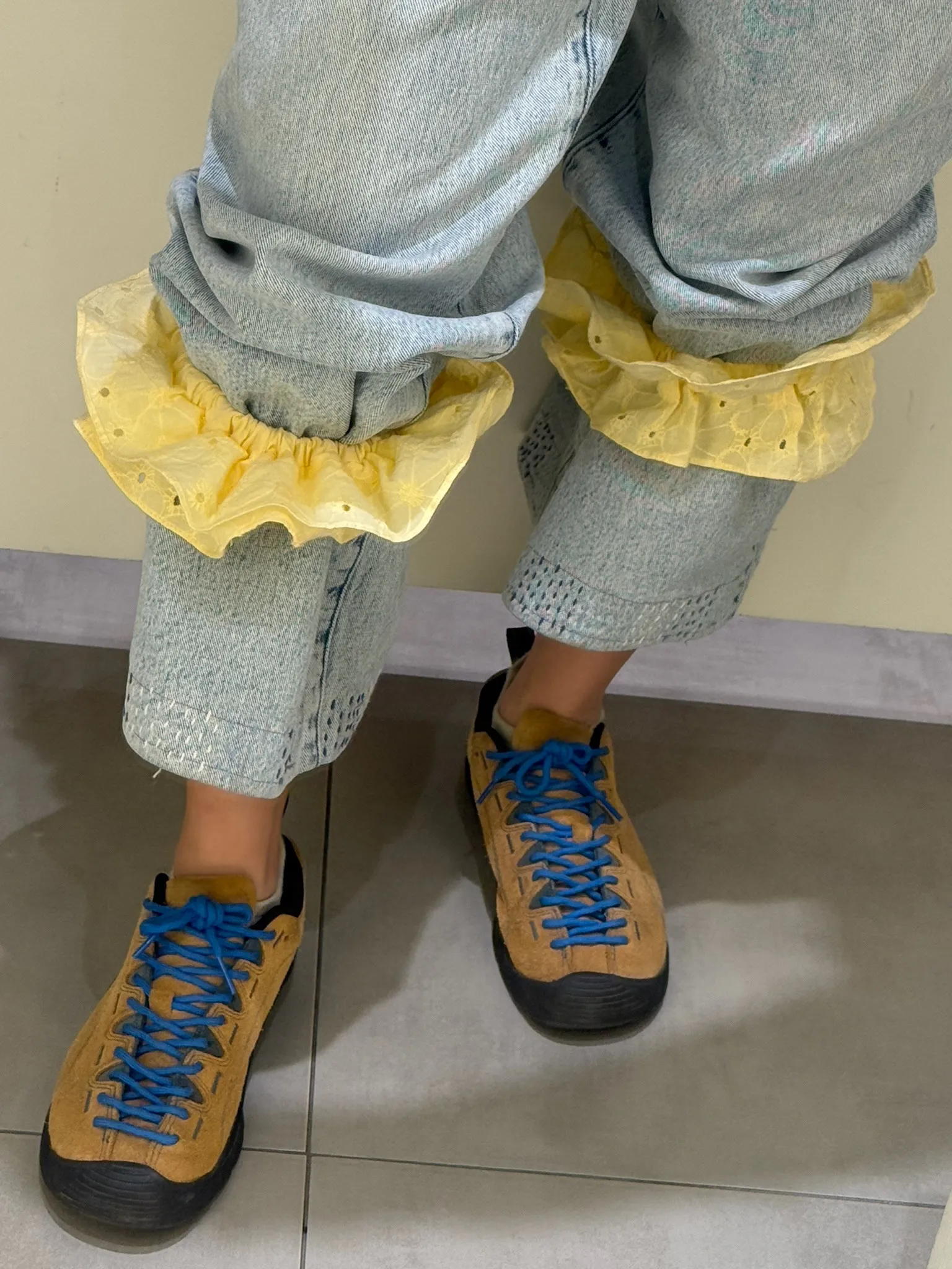 Yellow frilled HemHuggers gather pale blue denim trouser legs above tan walking shoes with blue laces.